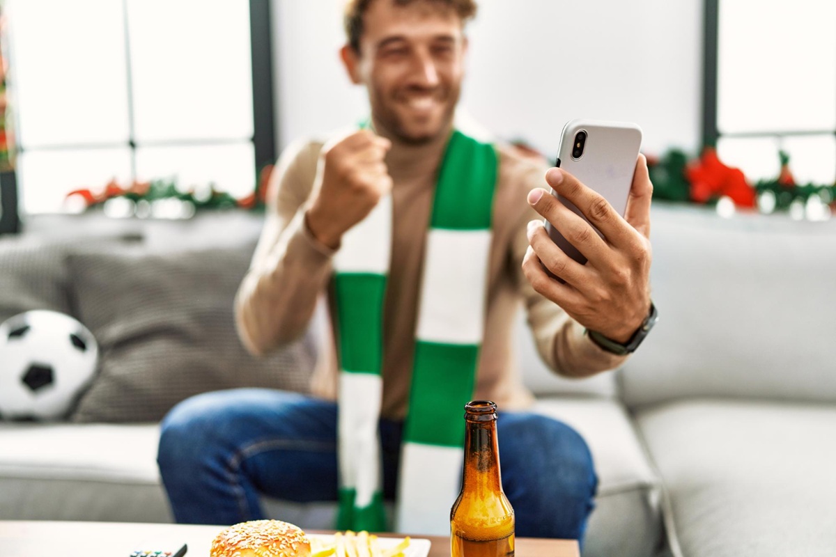 Homem assistindo jogo de futebol com celular fazendo apostas online enquanto bebe cerveja em casa