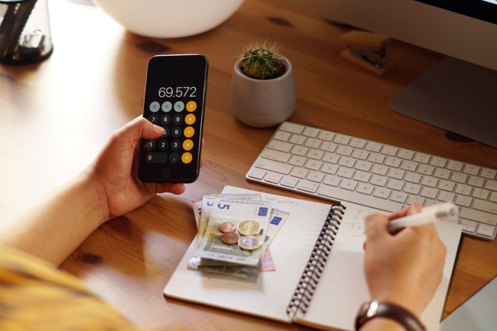 Pessoa utilizando calculadora e anotando em caderno com dinheiro ao lado, analisando gastos