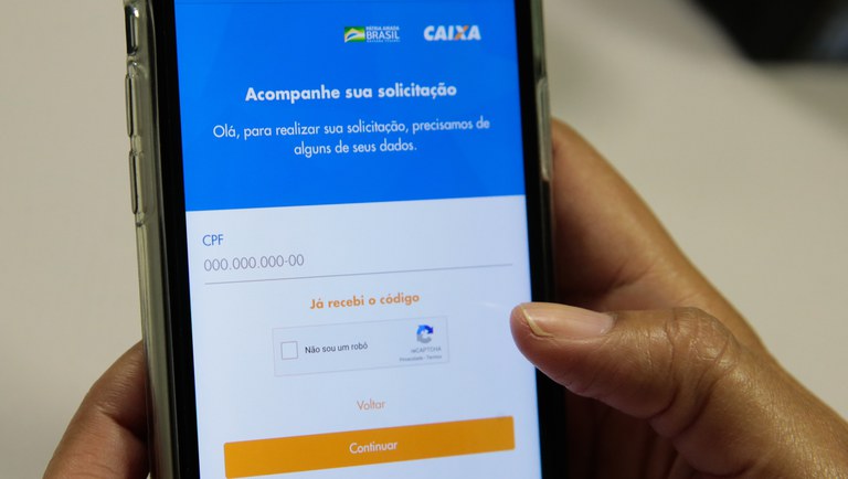 Mão segurando celular com tela de acompanhamento de solicitação da Caixa Econômica Federal do Auxilio Emergencial.