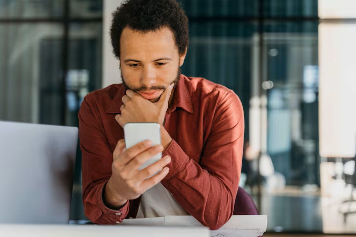 Homem de camisa bordô olhando para o celular, com expressão pensativa, em um ambiente de escritório.