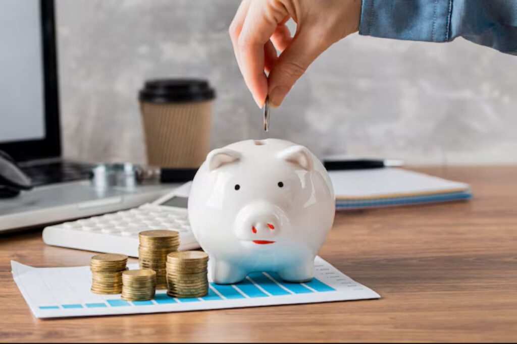 Pessoa colocando moeda em cofrinho de porcelana branca com pilhas de moedas ao lado, representando economia financeira.