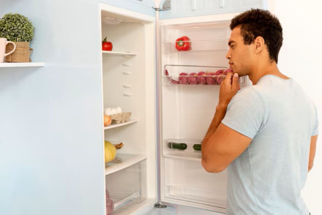 Homem pensativo observando geladeira aberta com poucos alimentos