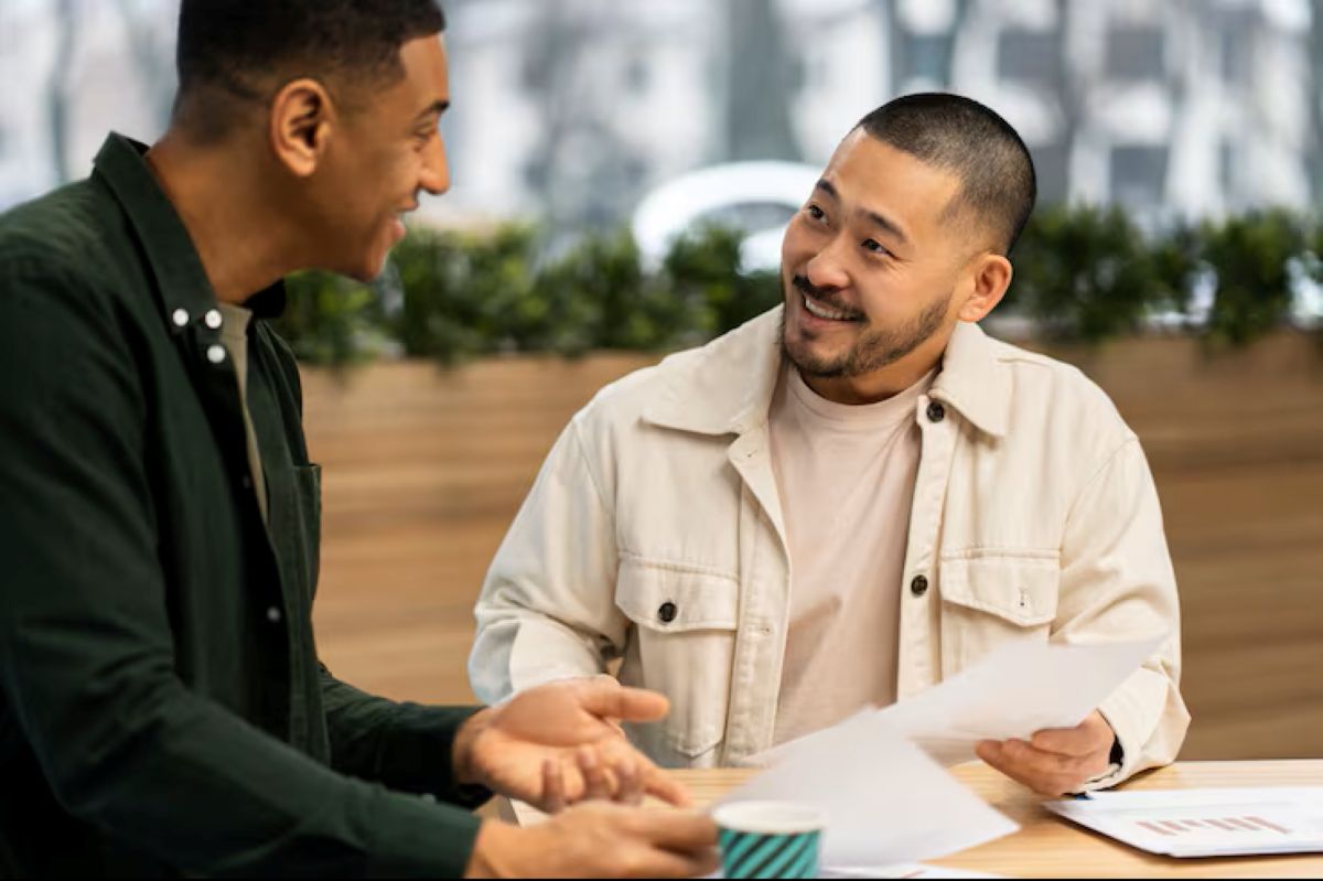 Dois amigos ou colegas conversando e sorrindo enquanto revisam documentos, simbolizando a importância de uma boa comunicação para preservar laços após tratar de finanças.