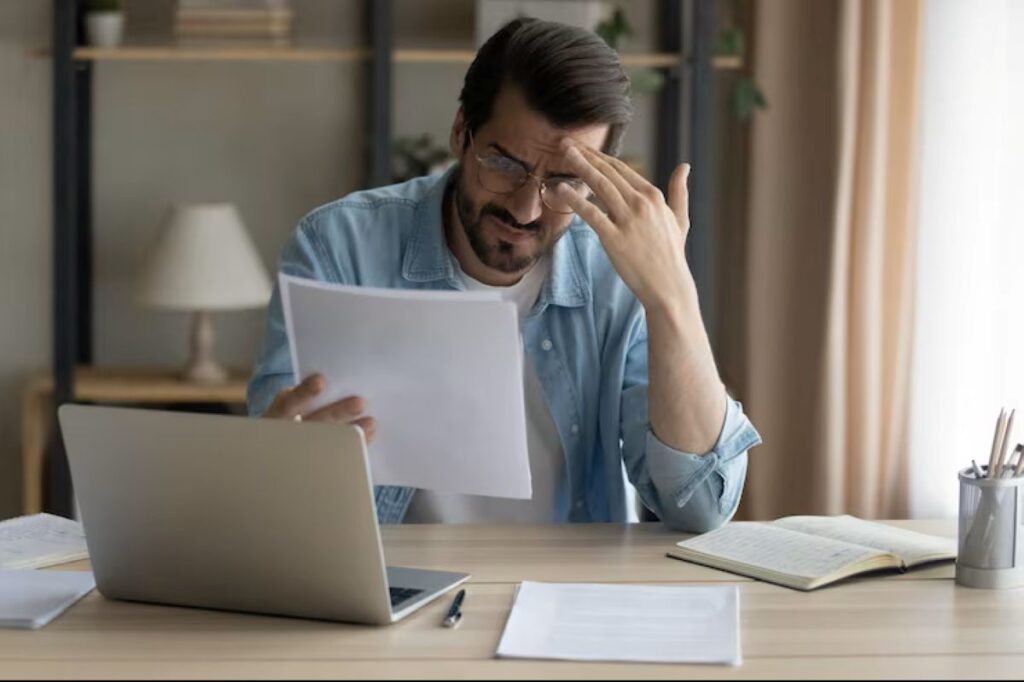 Homem com óculos, barba e camisa jeans, visivelmente frustrado, segura uma folha de papel e coloca a mão na testa, sentado à mesa de trabalho com um notebook, simbolizando a preocupação com uma dívida bancária.