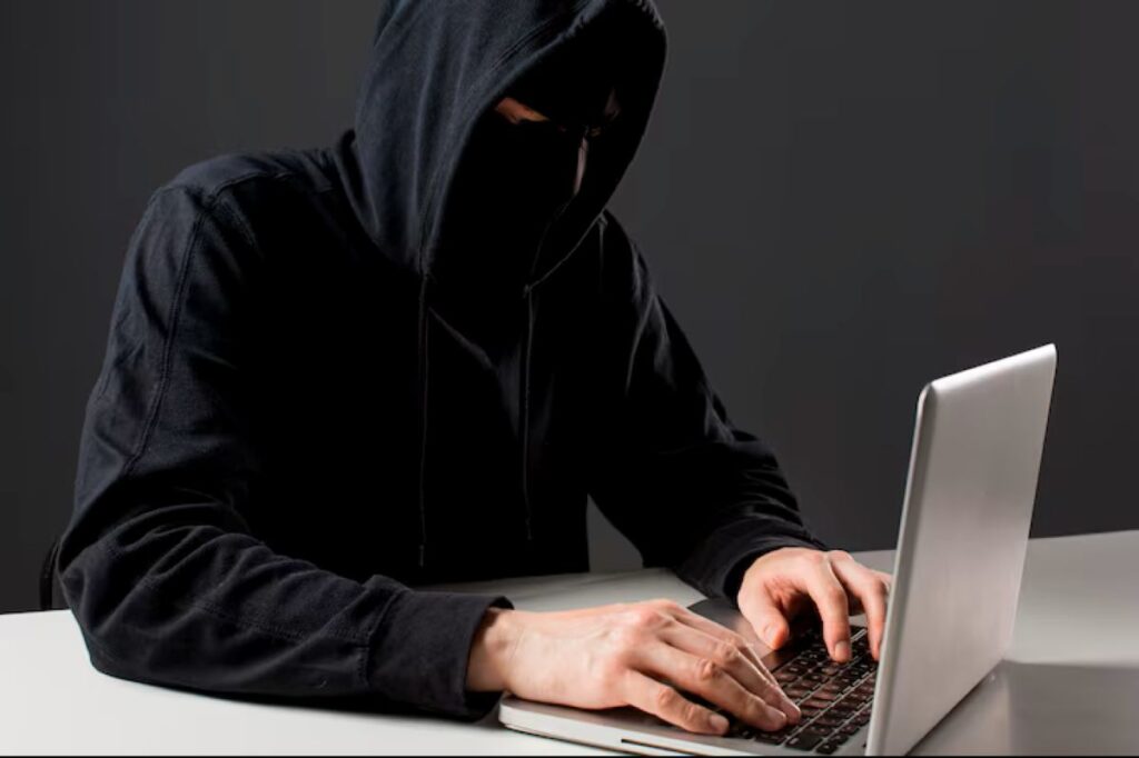 Pessoa encapuzada usando laptop para realizar atividades fraudulentas online.