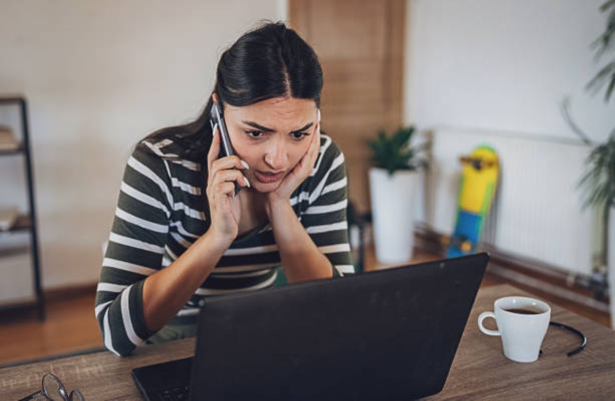 Mulher em casa, preocupada e conversando ao telefone enquanto usa um laptop, representando a angústia de ser vítima de fraude online.