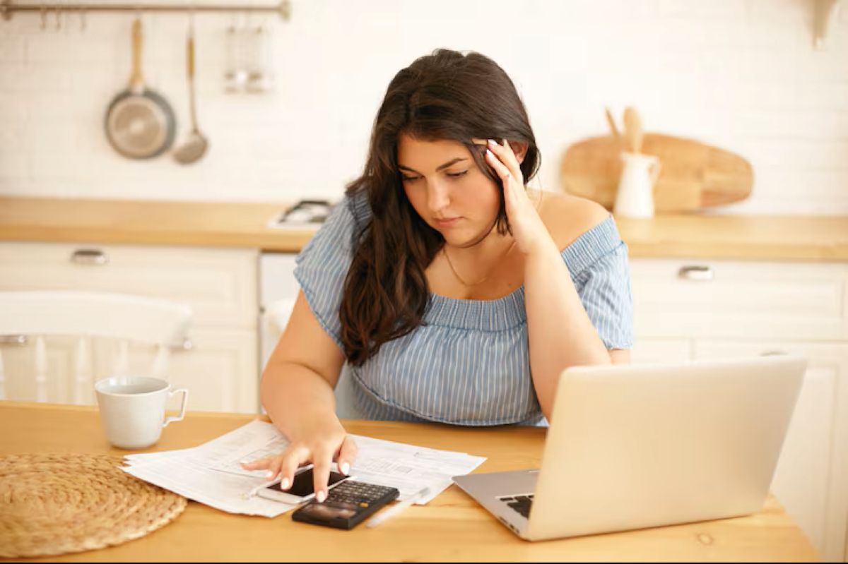 Mulher jovem com expressão preocupada, calculando dívidas em casa com celular, calculadora e laptop.