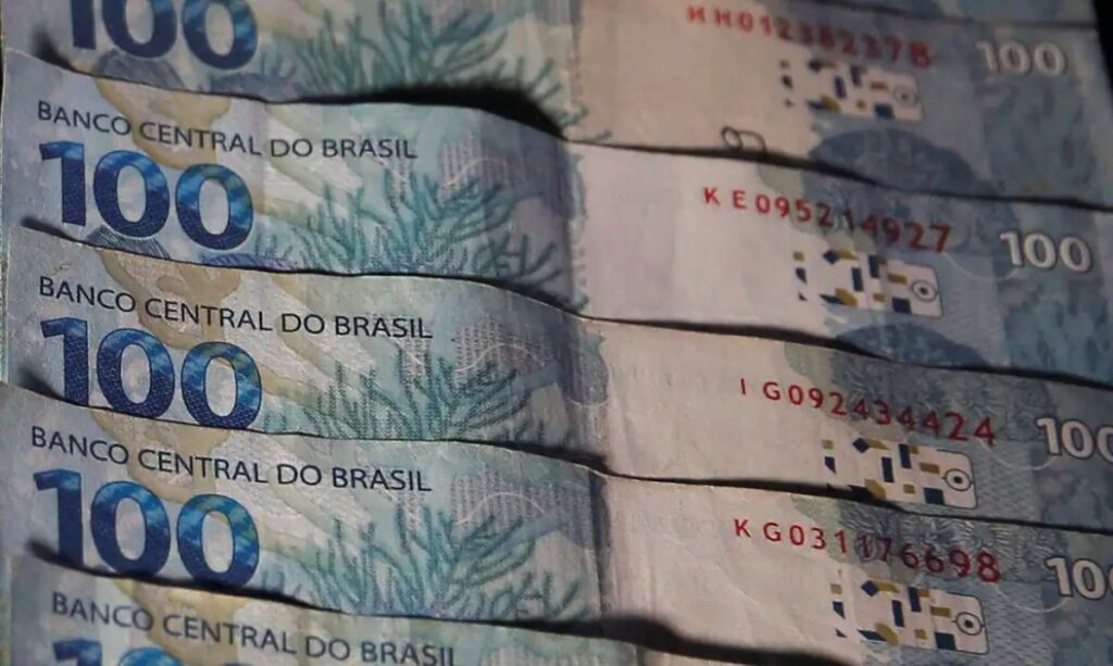Diversas cédulas de 100 reais alinhadas, com números de série visíveis e fundo azulado