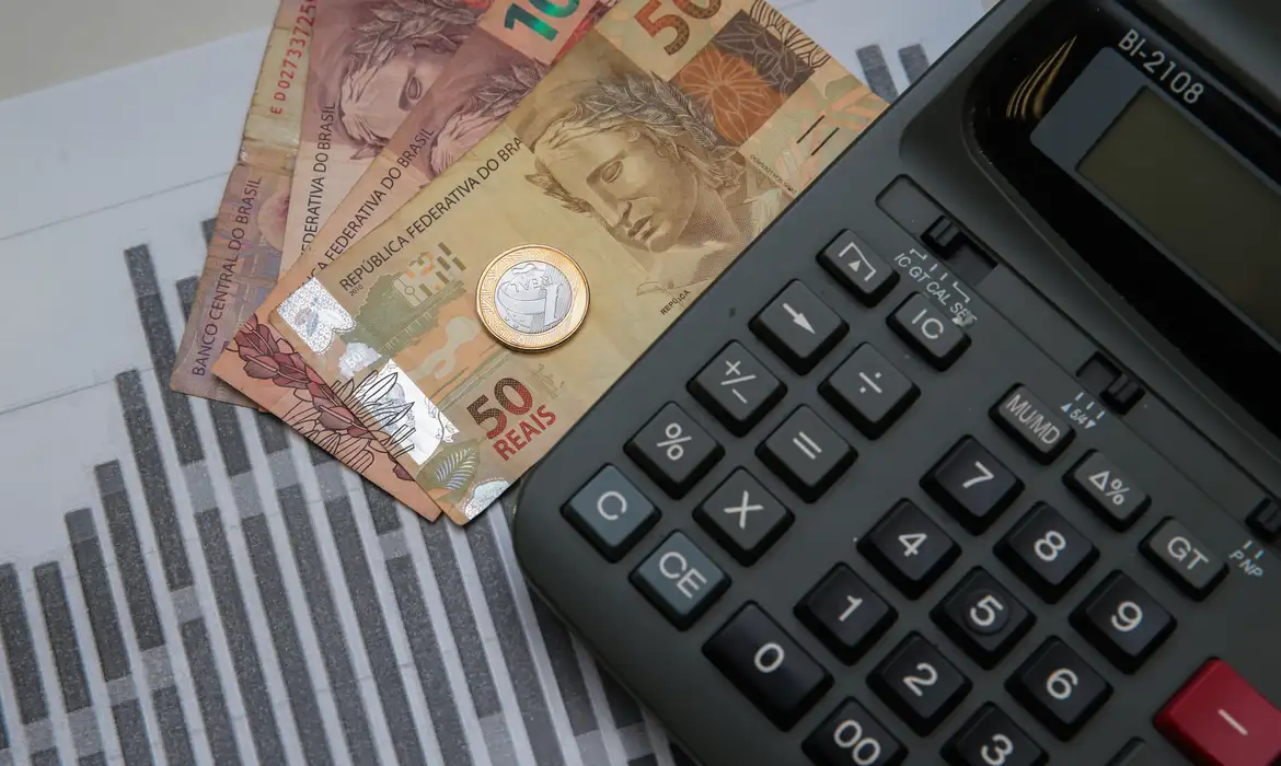 Notas de 100, 50 e 20 reais, moeda de 1 real e calculadora preta sobre gráfico de barras