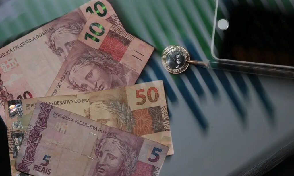 Notas de 5, 10 e 50 reais com moeda de 1 real e smartphone sobre mesa listrada