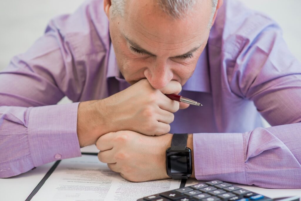 Imagem ilustrativa de homem preocupado com a mão no rosto diante de documentos e uma calculadora, representando o prejuízo causado pelos golpes financeiros