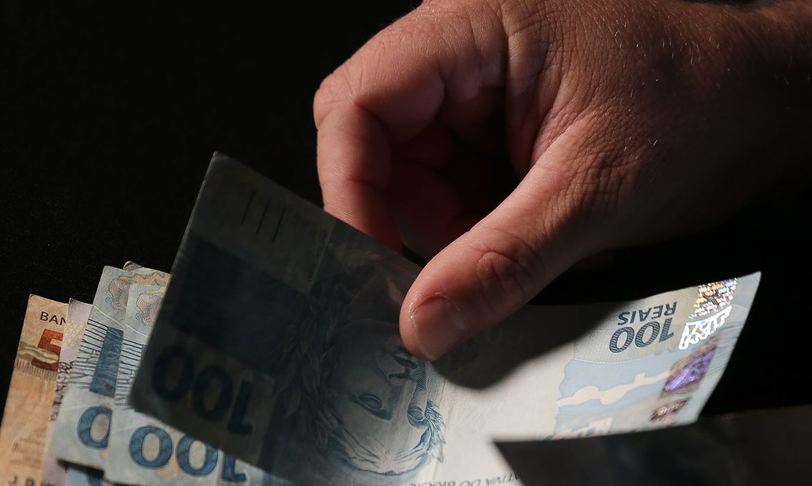 Mão masculina conta notas de real brasileiro, com cédulas de R$100 em primeiro plano