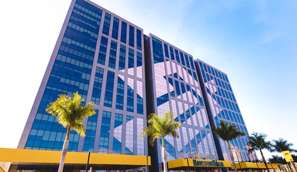 Fachada do edifício-sede do Banco do Brasil, prédio de vidro azul visto de baixo, com palmeiras à frente e marquise amarela com o nome da instituição na entrada.
