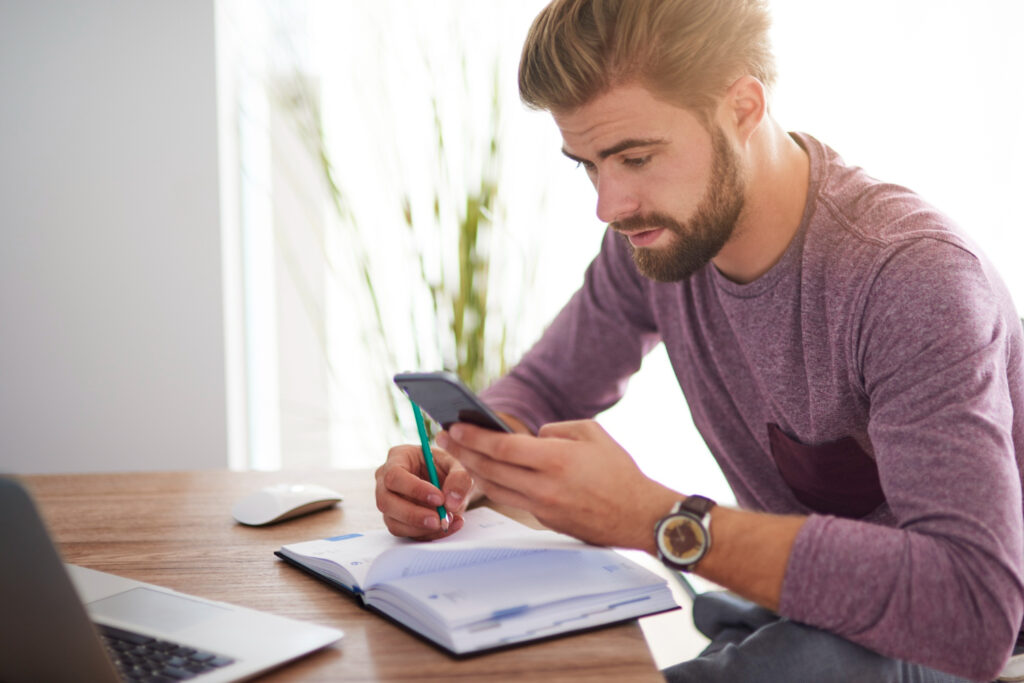 Imagem ilustrativa de homem usando o celular e anotando em um caderno ao lado do notebook, representando a consulta aos investimentos pelos canais digitais