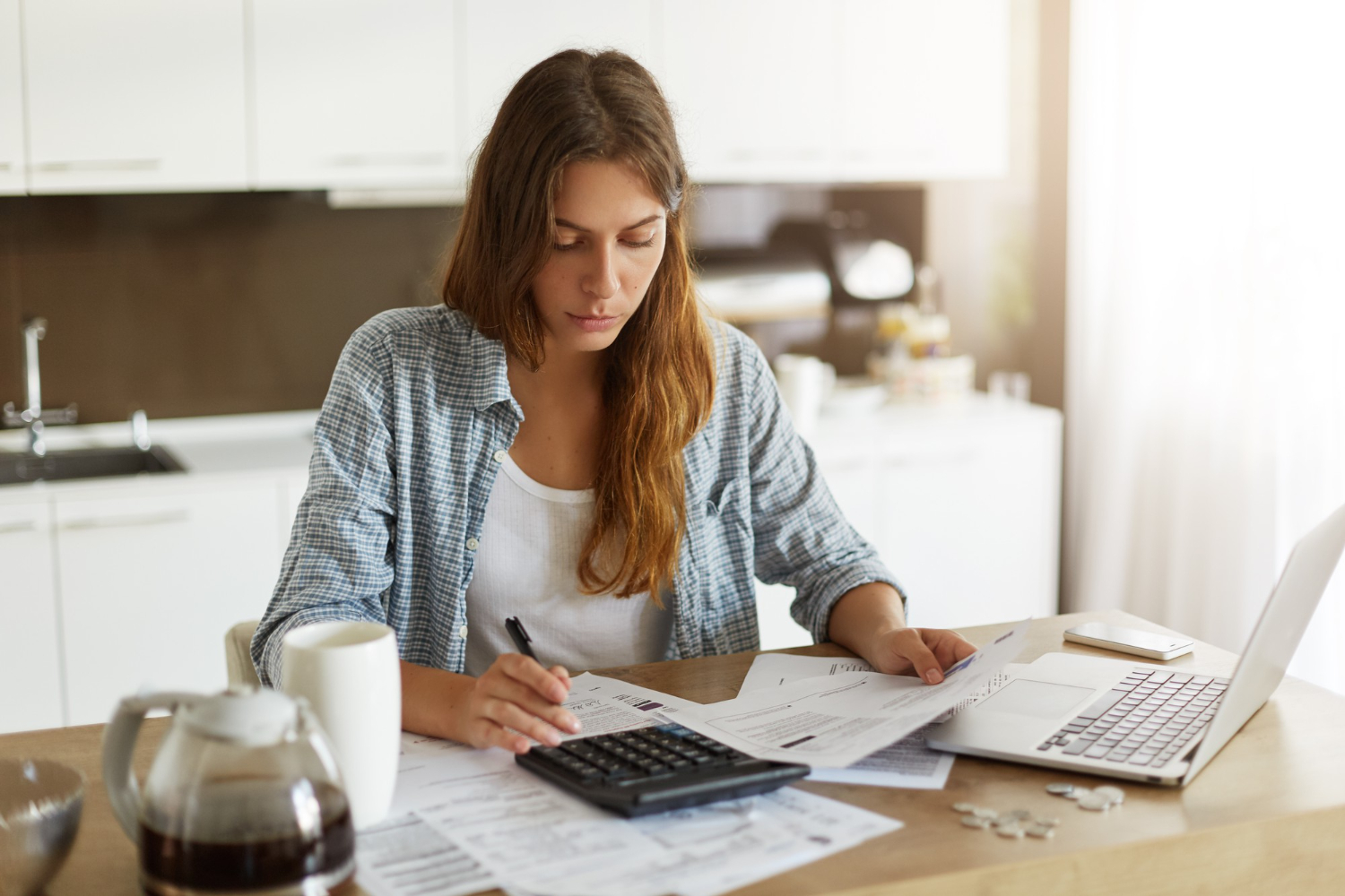 Imagem ilustrativa de mulher de camisa xadrez conferindo papéis com uma calculadora e um notebook sobre a mesa da cozinha, referência à revisão do orçamento para recomeçar o planejamento