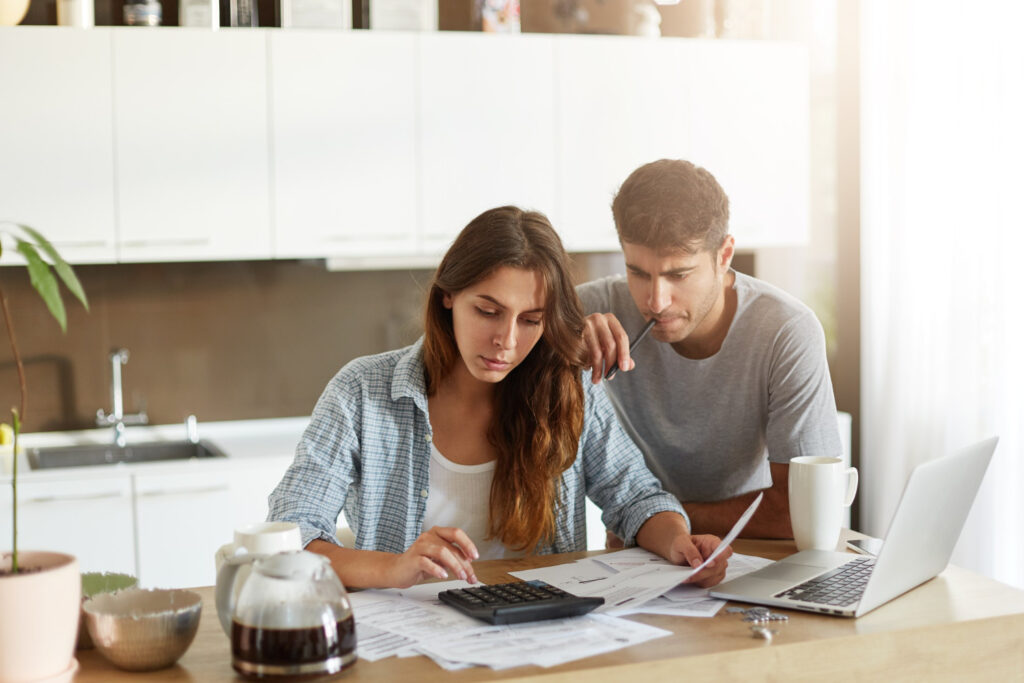 Imagem ilustrativa de casal na cozinha conferindo contas com calculadora, papéis e notebook sobre a mesa, em referência ao cálculo da divisão de despesas entre os dois
