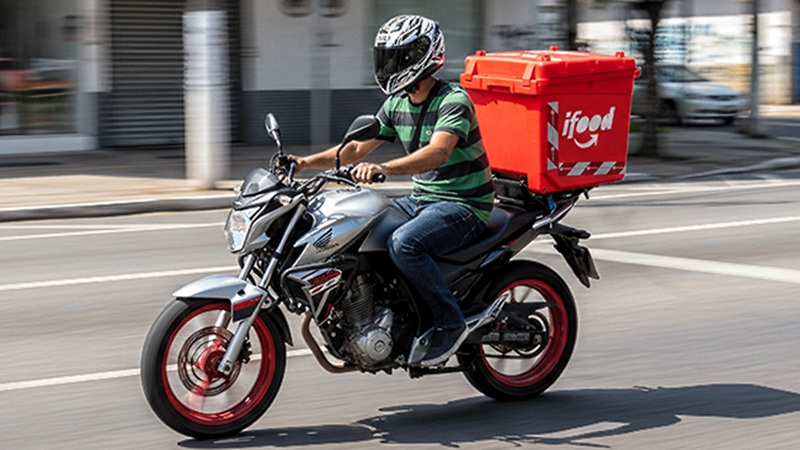 Entregador de capacete pilotando uma moto com o baú vermelho do iFood acoplado na traseira, em movimento por uma avenida, referência ao público que atende aos requisitos da linha de crédito