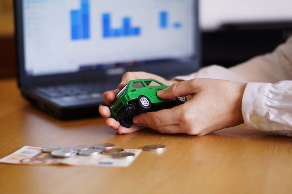 Imagem ilustrativa de mãos segurando a miniatura de um carro sobre a mesa, com moedas, cédula e um notebook exibindo gráficos ao fundo, cena ligada à busca por financiamento de veículo.