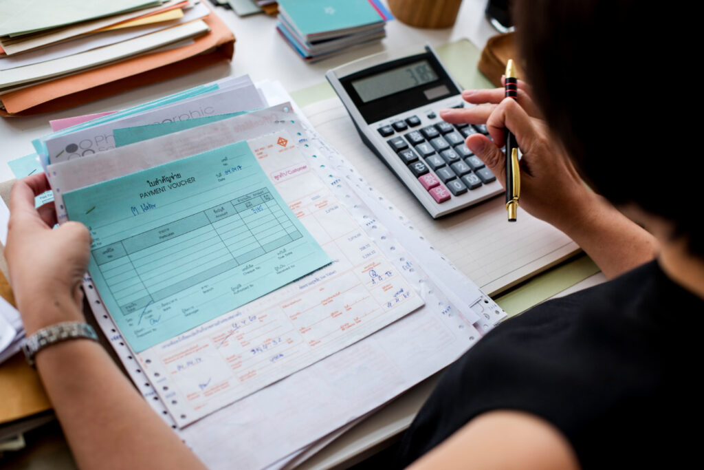 Imagem ilustrativa de mulher conferindo uma pilha de comprovantes de pagamento com o auxílio de uma calculadora de mesa, em referência à divisão do orçamento mensal.