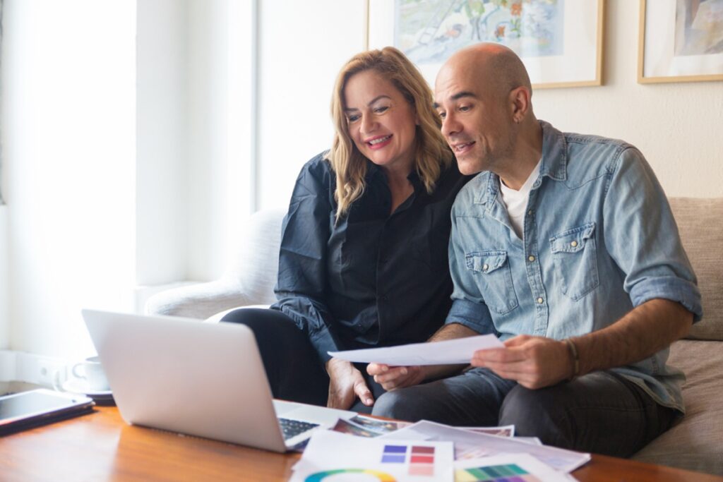 Imagem ilustrativa de casal sentado no sofá diante de um notebook e de papéis com anotações, em referência às formas de organizar as finanças da vida a dois