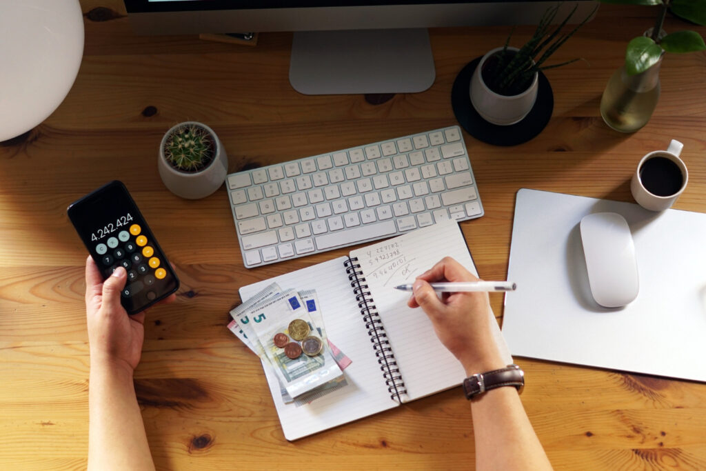 Imagem ilustrativa de mesa vista de cima com pessoa anotando cálculos em caderno, celular aberto na calculadora, cédulas e moedas ao lado do teclado, tema da matéria sobre organização financeira com um salário mínimo.