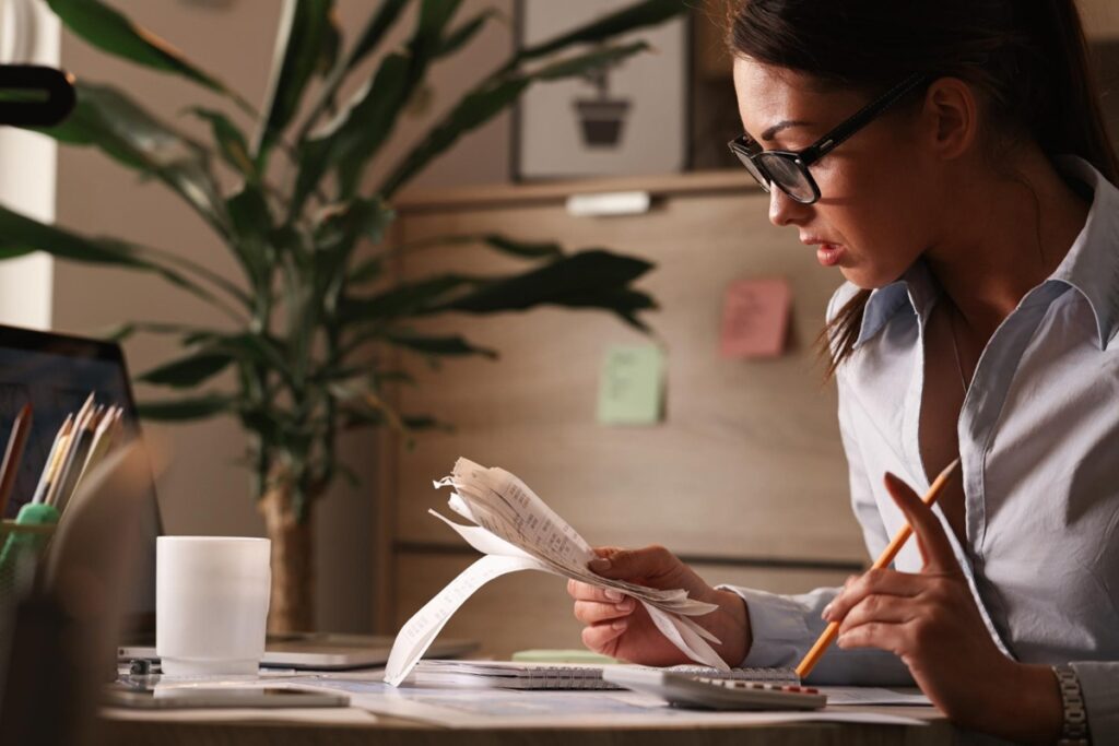 Imagem ilustrativa de mulher de óculos conferindo uma sequência de comprovantes em papel com lápis na mão e calculadora sobre a mesa, referência à definição de metas financeiras possíveis de cumprir