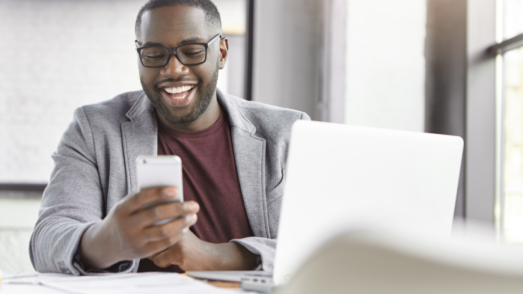 Jovem sorrindo enquanto usa celular e notebook em ambiente moderno.