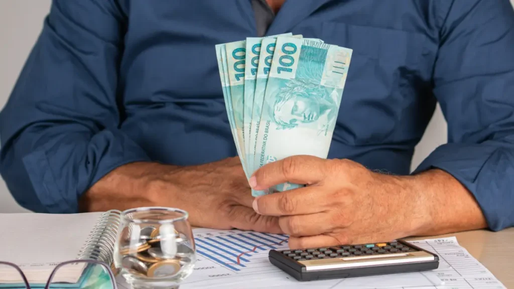 Homem segura notas de R$100 ao lado de calculadora e moedas.