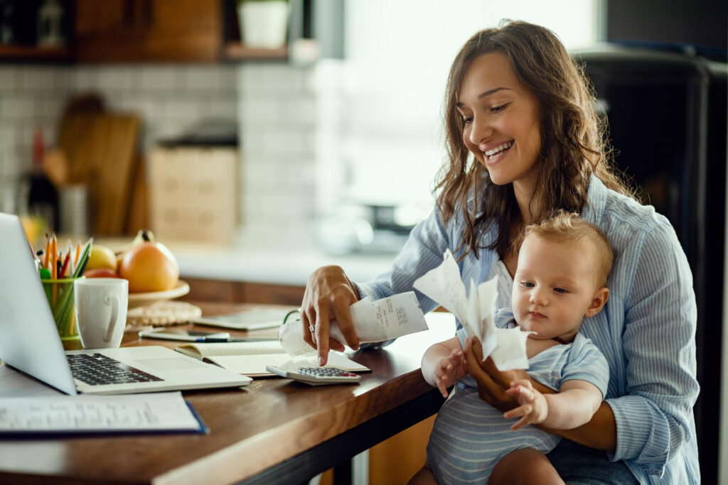 Imagem ilustrativa: mulher com bebê no colo confere recibos e calculadora, cena ligada aos gastos do Dia das Crianças
