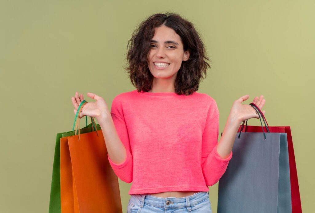 Imagem ilustrativa de mulher sorridente segurando várias sacolas de papel coloridas em cada mão, sobre fundo verde.