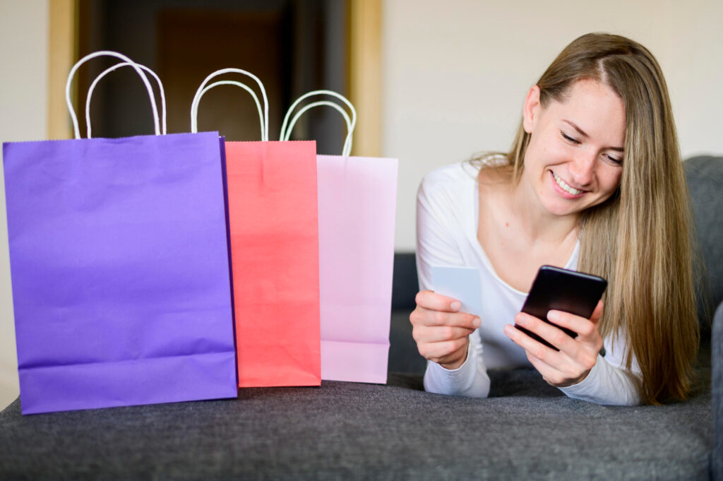 Imagem ilustrativa de mulher deitada no sofá com celular na mão, ao lado de sacolas de compras coloridas.