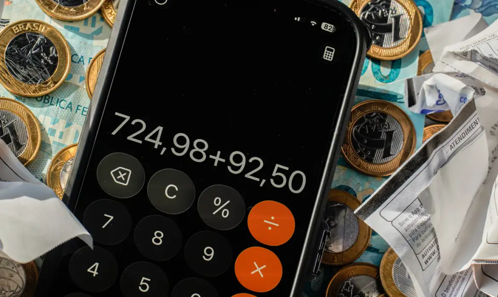 Imagem ilustrativa de celular com a calculadora aberta somando valores, apoiado sobre moedas, notas de real e comprovantes amassados, representando o efeito da inflação nas contas da casa.