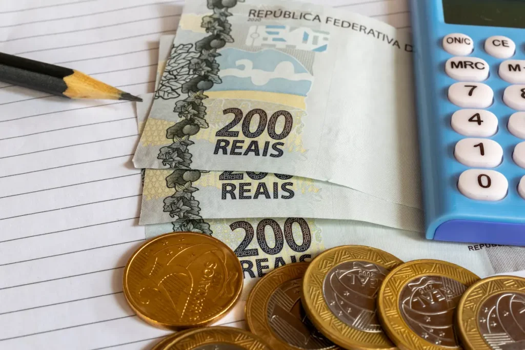 Notas e moedas de real ao lado de calculadora e caderno para controle financeiro.