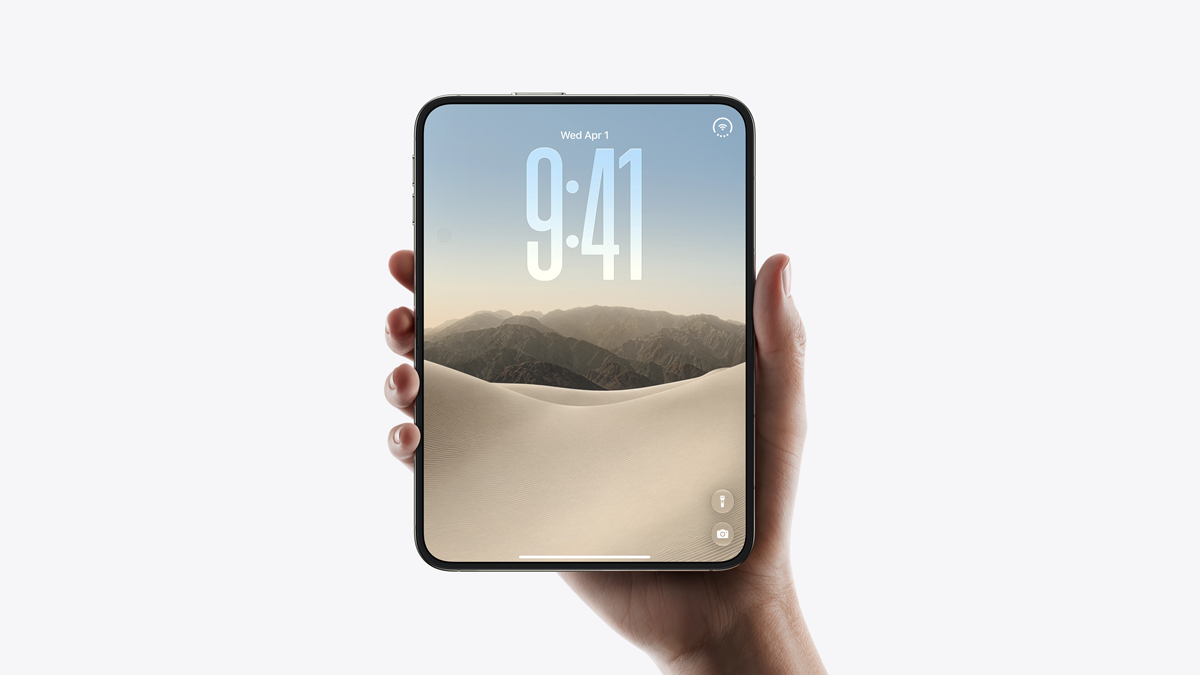 Mão segurando o novo iPhone Duo fechado, com a tela externa exibindo o relógio sobre uma paisagem de dunas